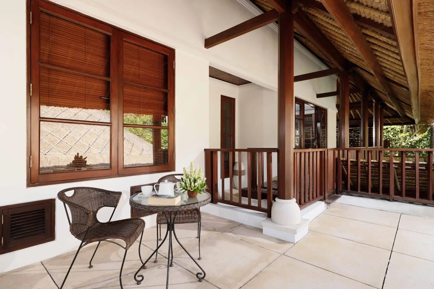 Cozy wicker chair and round table with tea set on a Balinese-style veranda, featuring thatched roof, wooden shutters, and tropical garden views. - Bali Villas