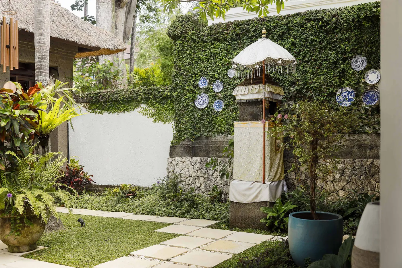 Cozy wicker chair and round table with tea set on a Balinese-style veranda, featuring thatched roof, wooden shutters, and tropical garden views. - Bali Villas