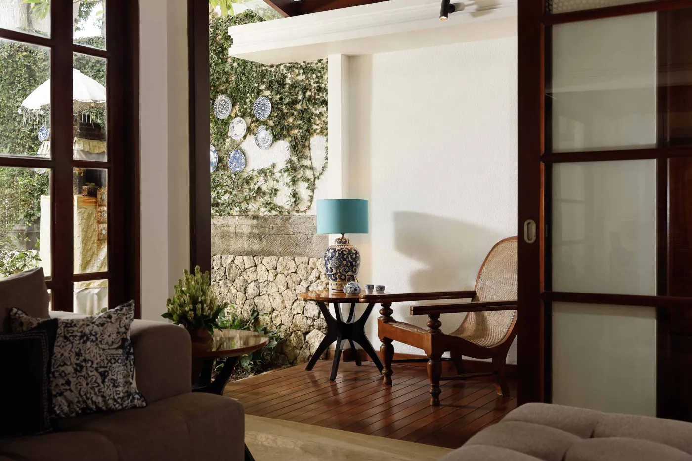 Cozy wicker chair and round table with tea set on a Balinese-style veranda, featuring thatched roof, wooden shutters, and tropical garden views. - Bali Villas