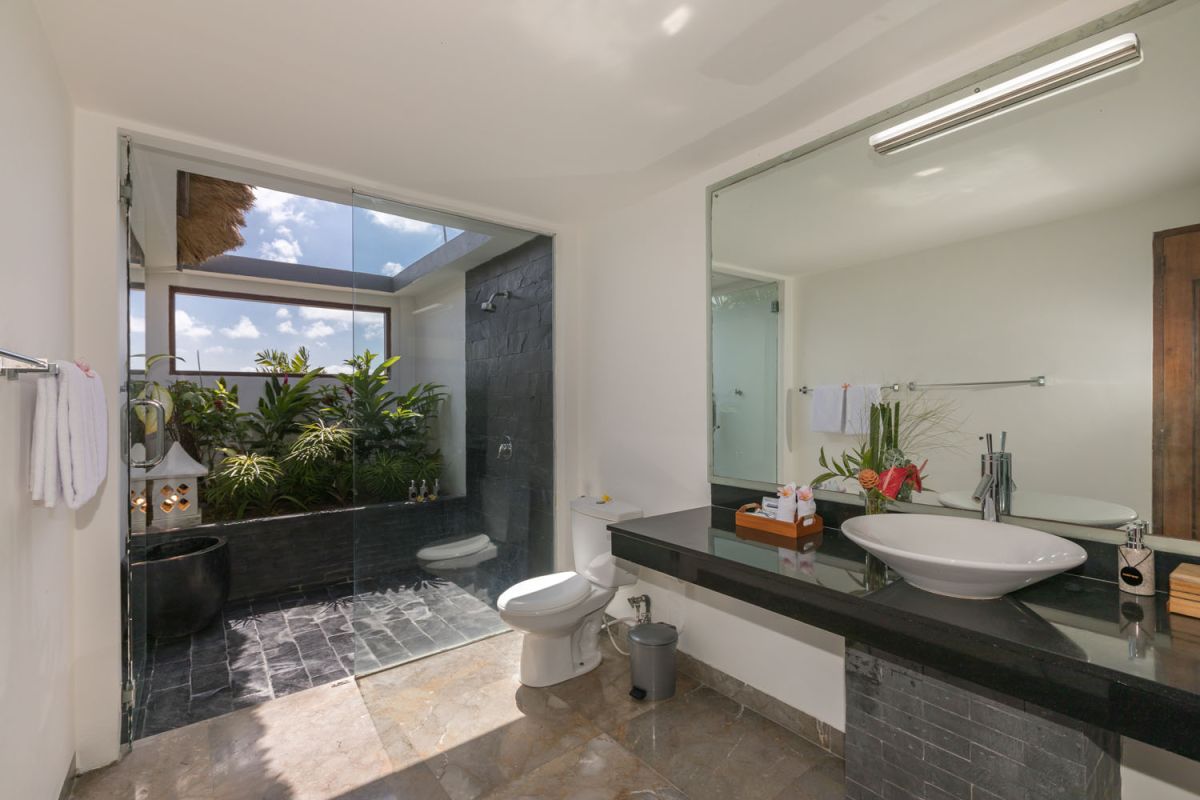 Spacious Balinese-style bathroom with tropical plants, open glass shower, white vanity, and skylight. - Bali Villas