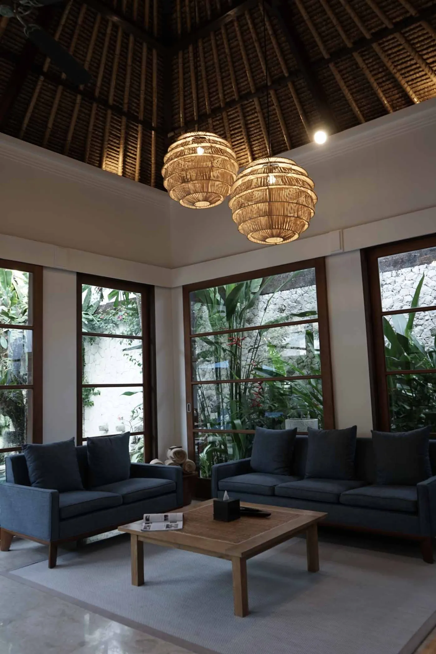 Balinese villa interior featuring a woven thatched hanging lamp, open white kitchen, and vibrant rooster wall art over a wooden dining table. - Bali Villas