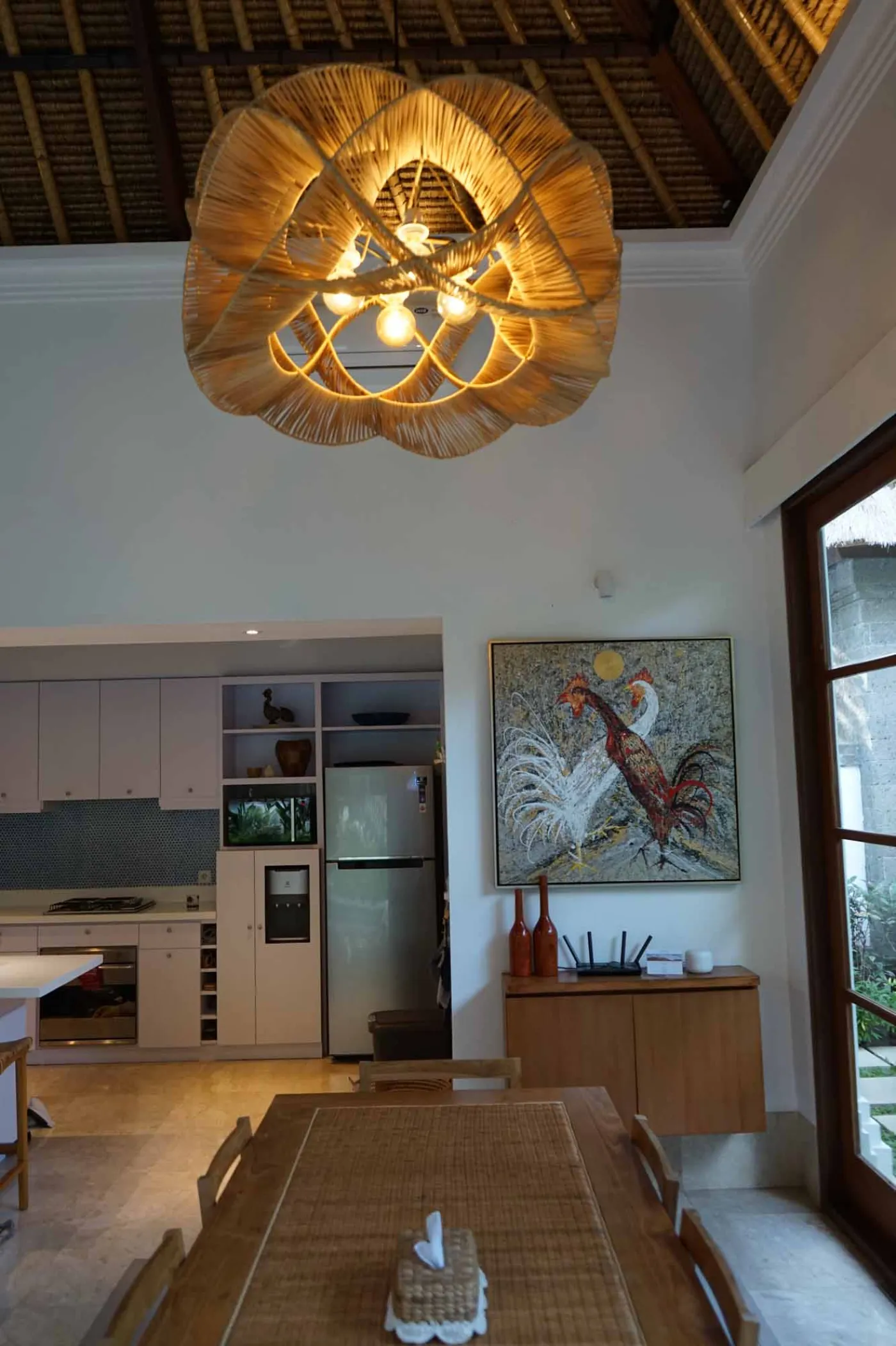 Balinese villa interior featuring a woven thatched hanging lamp, open white kitchen, and vibrant rooster wall art over a wooden dining table. - Bali Villas