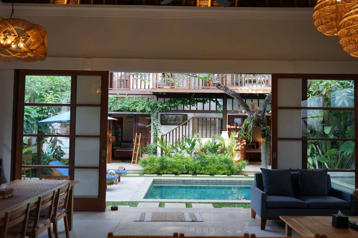 Balinese villa interior featuring a woven thatched hanging lamp, open white kitchen, and vibrant rooster wall art over a wooden dining table. - Bali Villas