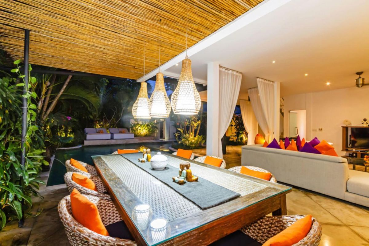 Modern Balinese-style kitchen with bamboo pendant lights, concrete island bar, white cabinets, tropical flower vase, and wooden statues. - Bali Villas