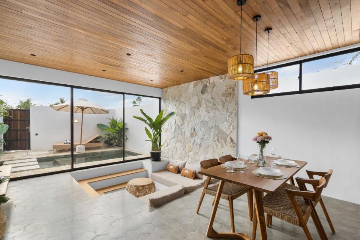 Modern tropical kitchen-dining area with wooden cabinets, rattan pendant lights, woven wall decor, and a set table under a beamed ceiling. - Bali Villas