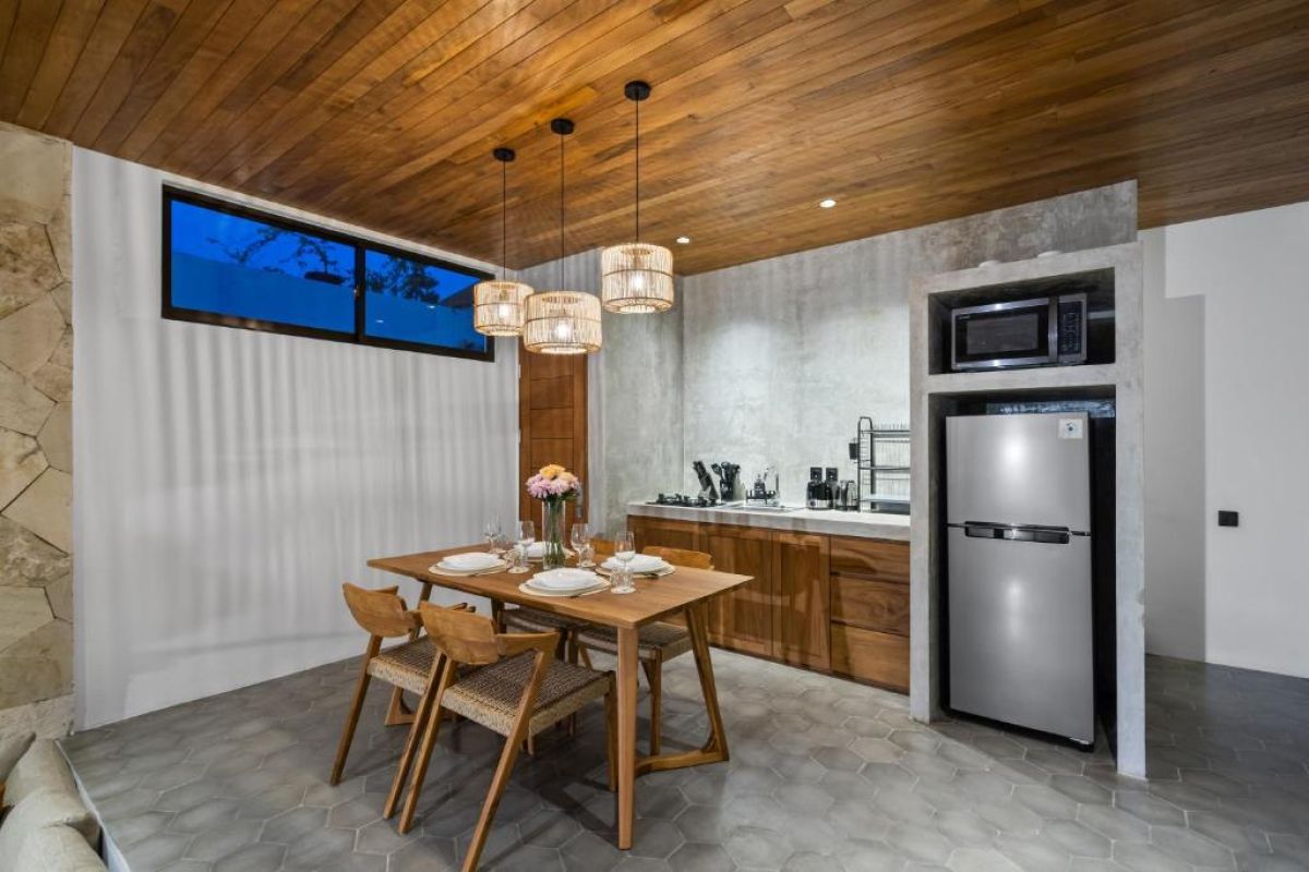 Modern tropical kitchen-dining area with wooden cabinets, rattan pendant lights, woven wall decor, and a set table under a beamed ceiling. - Bali Villas