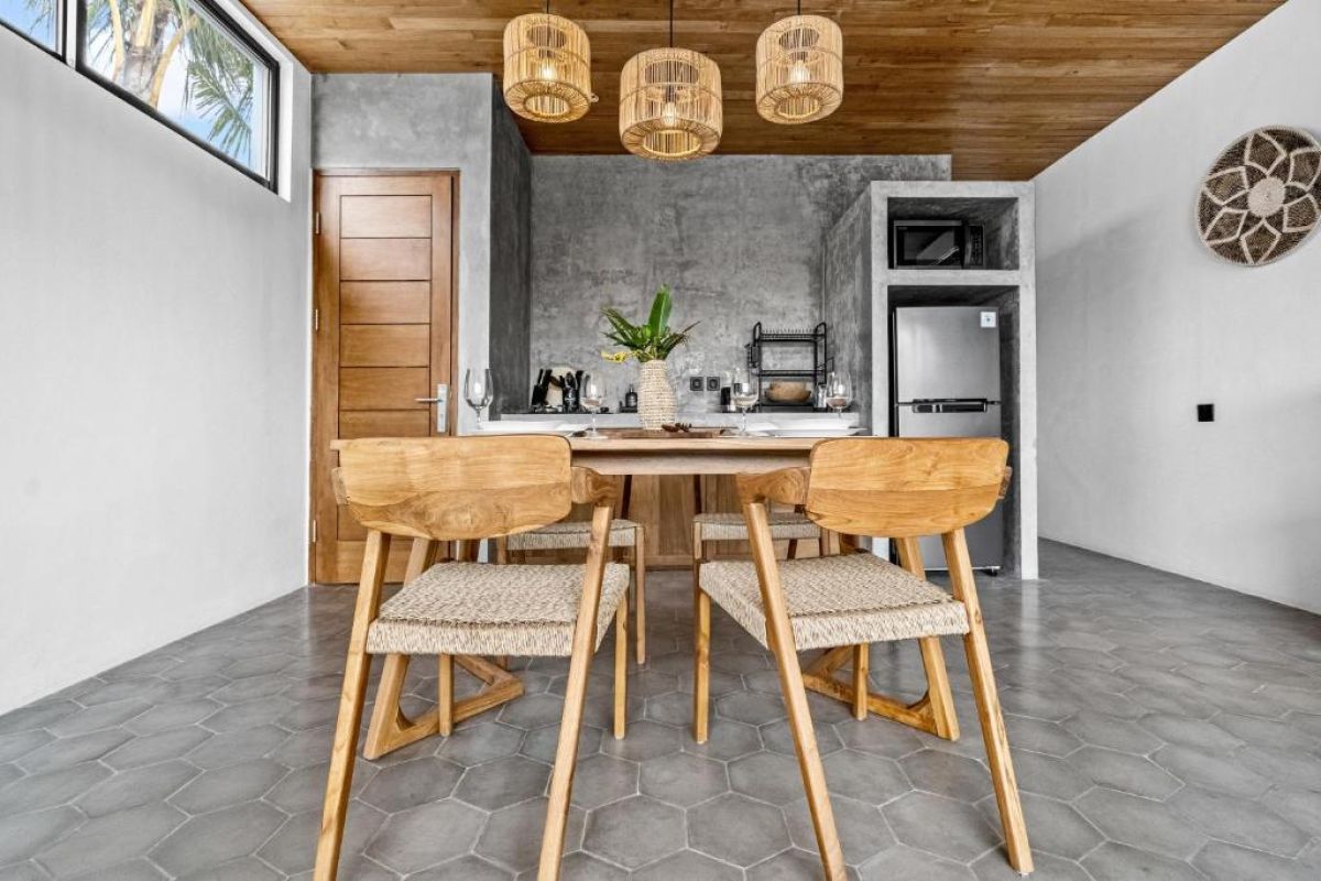 Modern tropical kitchen-dining area with wooden cabinets, rattan pendant lights, woven wall decor, and a set table under a beamed ceiling. - Bali Villas
