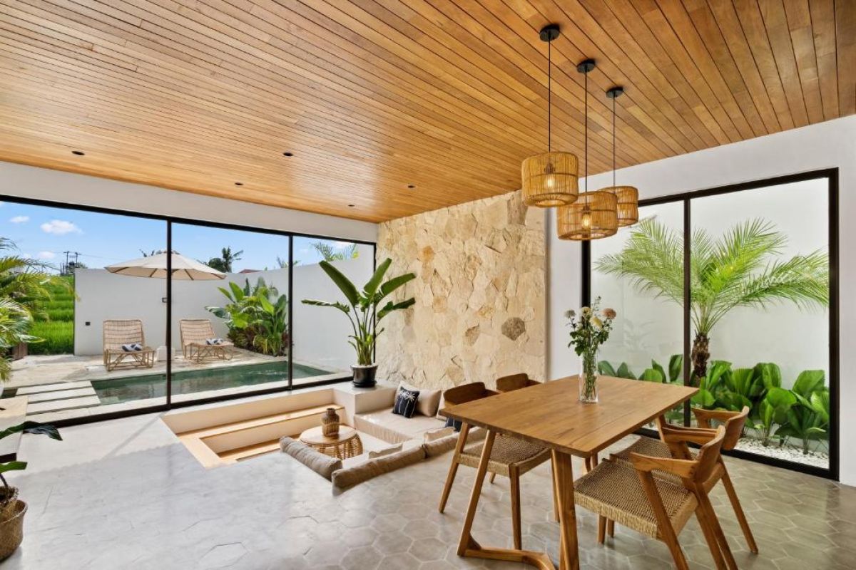 Modern tropical kitchen-dining area with wooden cabinets, rattan pendant lights, woven wall decor, and a set table under a beamed ceiling. - Bali Villas