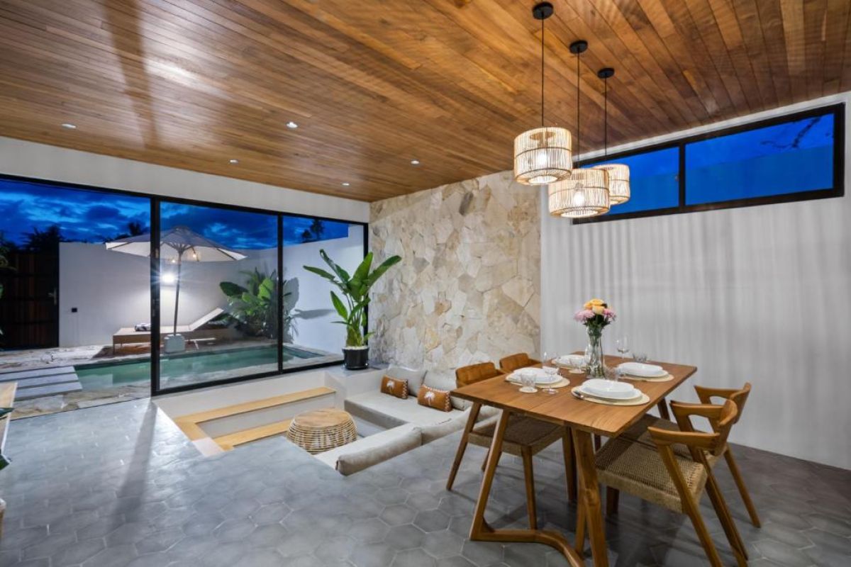 Modern tropical kitchen-dining area with wooden cabinets, rattan pendant lights, woven wall decor, and a set table under a beamed ceiling. - Bali Villas