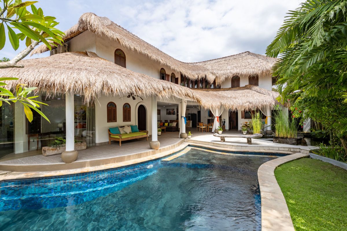 Tropical Balinese villa with curved thatched roof, white stucco walls, infinity pool, and lush palm garden. - Bali Villas