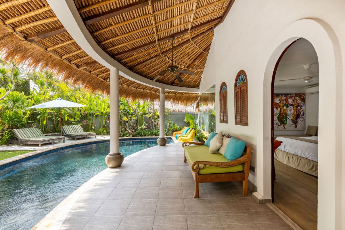 Spacious tropical villa veranda with curved thatched roof, wooden dining table and chairs, lush greenery, hanging drapes, and a serene lily pond. - Bali Villas