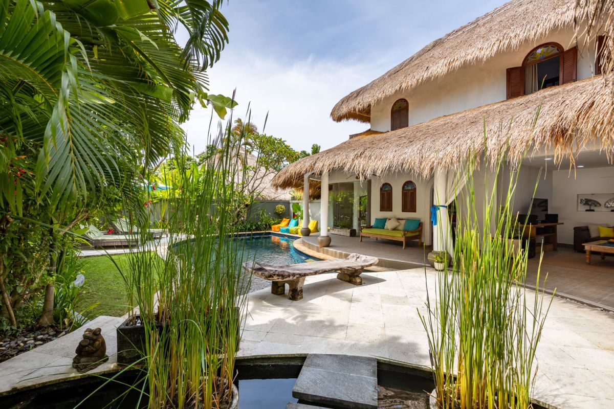 Spacious tropical villa veranda with curved thatched roof, wooden dining table and chairs, lush greenery, hanging drapes, and a serene lily pond. - Bali Villas