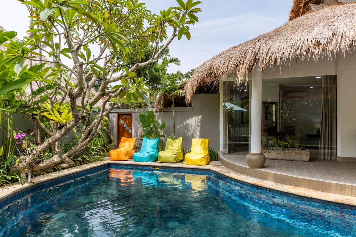 Spacious tropical villa veranda with curved thatched roof, wooden dining table and chairs, lush greenery, hanging drapes, and a serene lily pond. - Bali Villas