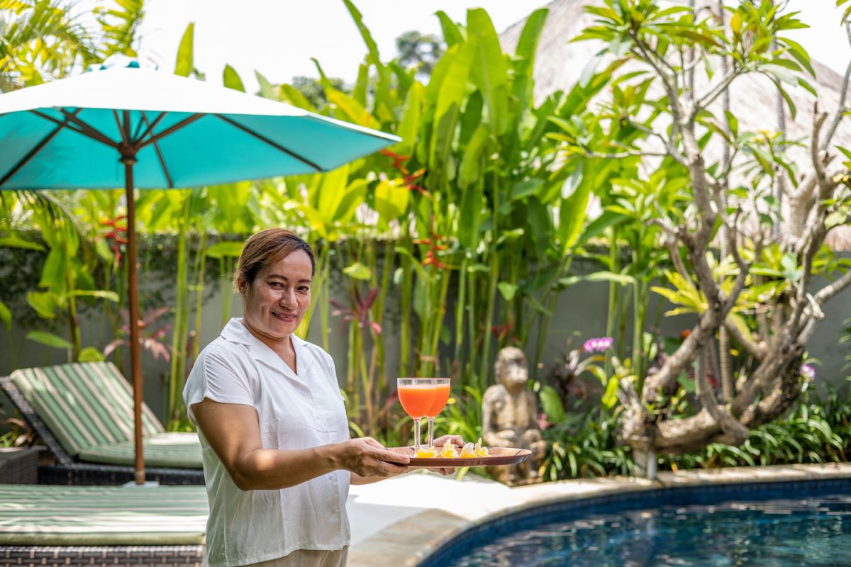 Spacious tropical villa veranda with curved thatched roof, wooden dining table and chairs, lush greenery, hanging drapes, and a serene lily pond. - Bali Villas