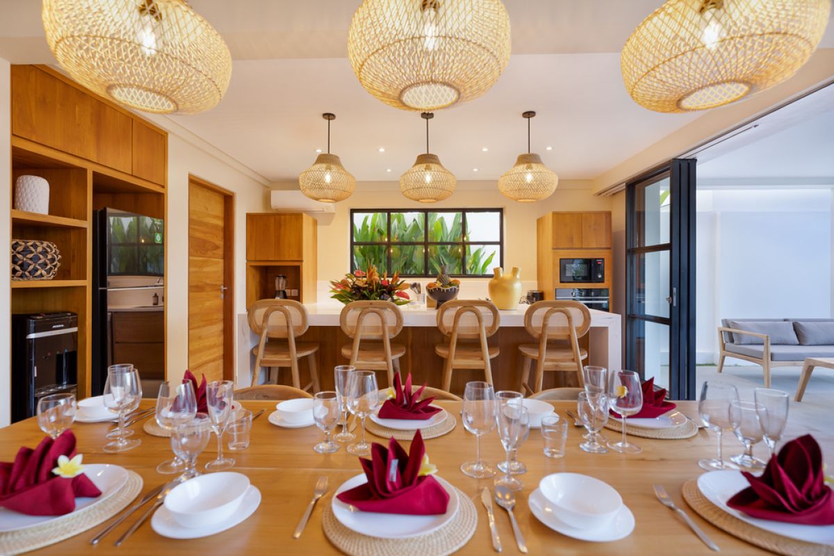 Elegant open-plan tropical kitchen with wooden cabinets, bamboo pendant lights, set dining table, and sliding glass doors to outdoor patio. - Bali Villas