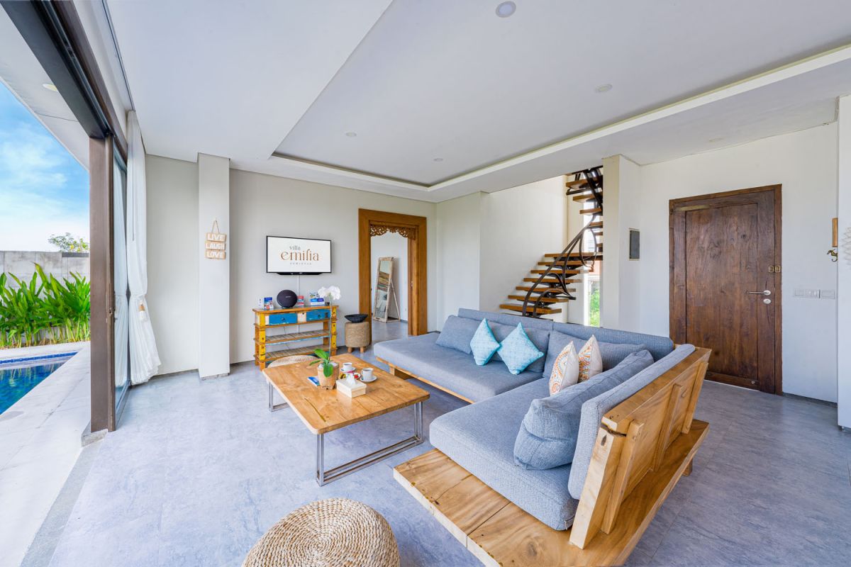 Spacious modern tropical dining room with wooden table, benches, and chairs, tropical flower centerpiece, open kitchen, and large windows overlooking lush greenery. - Bali Villas