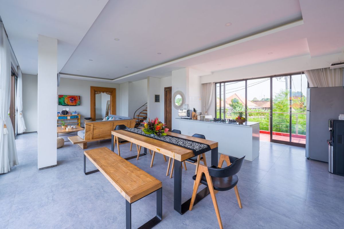 Spacious modern tropical dining room with wooden table, benches, and chairs, tropical flower centerpiece, open kitchen, and large windows overlooking lush greenery. - Bali Villas