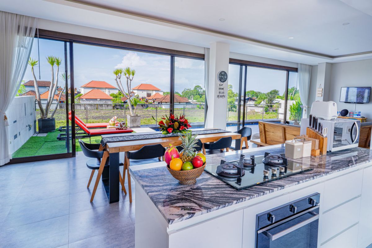 Spacious modern tropical dining room with wooden table, benches, and chairs, tropical flower centerpiece, open kitchen, and large windows overlooking lush greenery. - Bali Villas