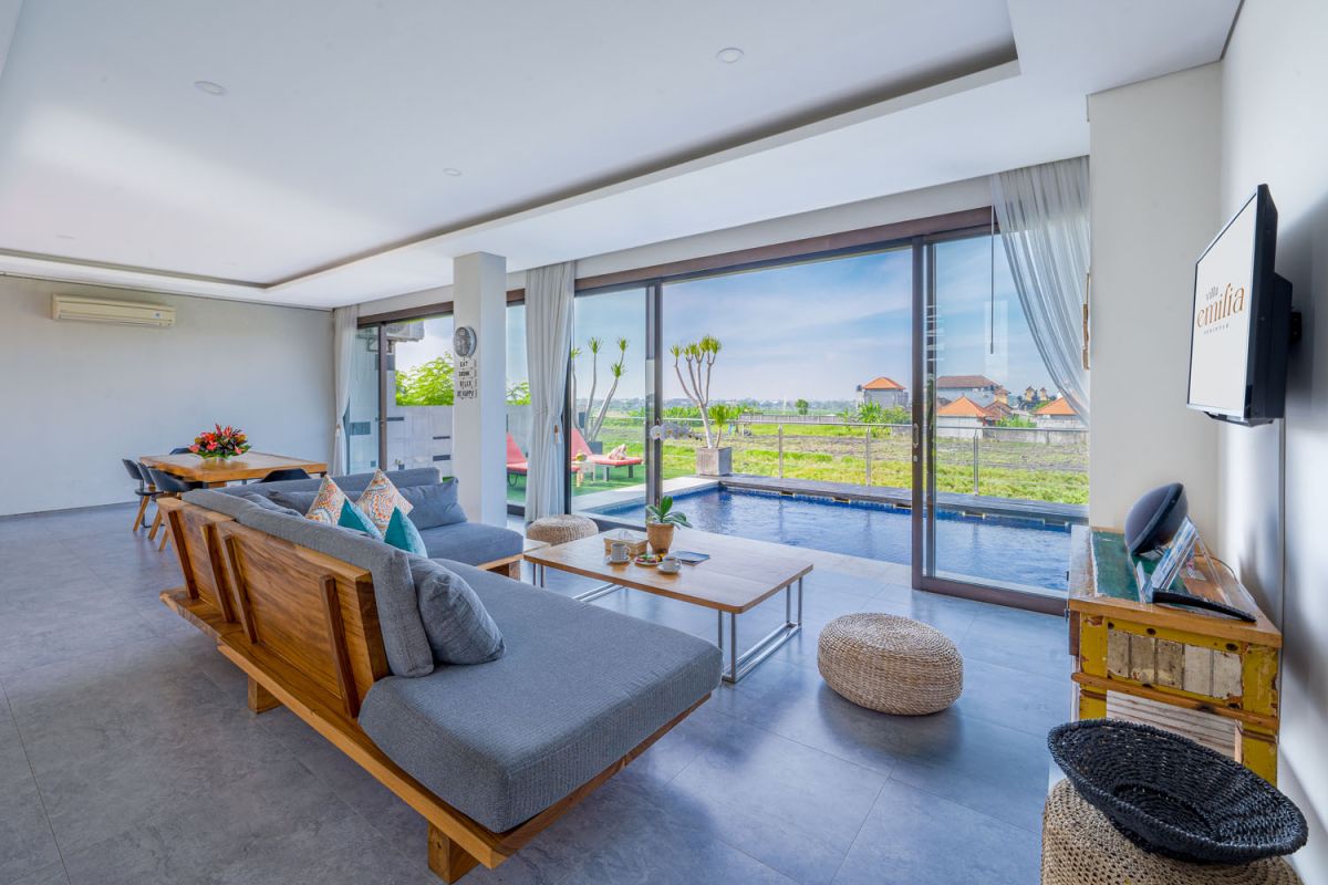 Spacious modern tropical dining room with wooden table, benches, and chairs, tropical flower centerpiece, open kitchen, and large windows overlooking lush greenery. - Bali Villas