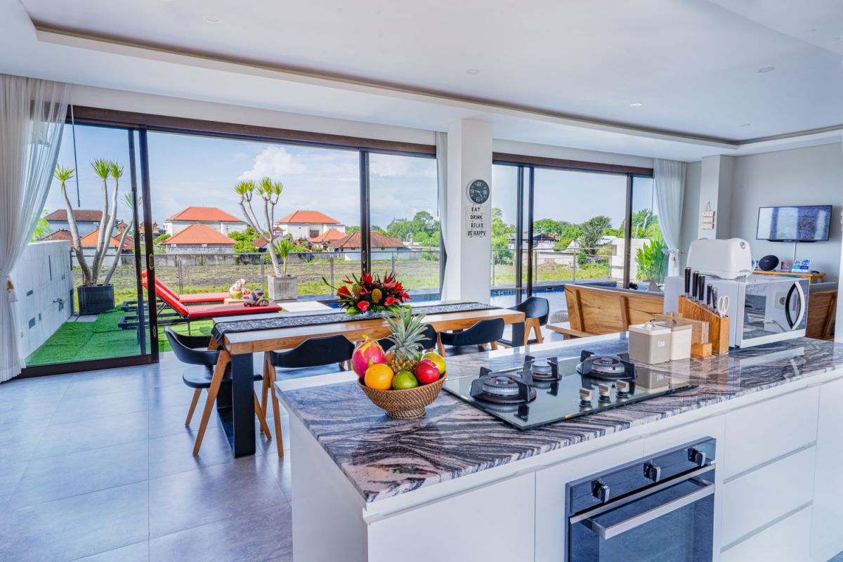 Spacious modern tropical dining room with wooden table, benches, and chairs, tropical flower centerpiece, open kitchen, and large windows overlooking lush greenery. - Bali Villas