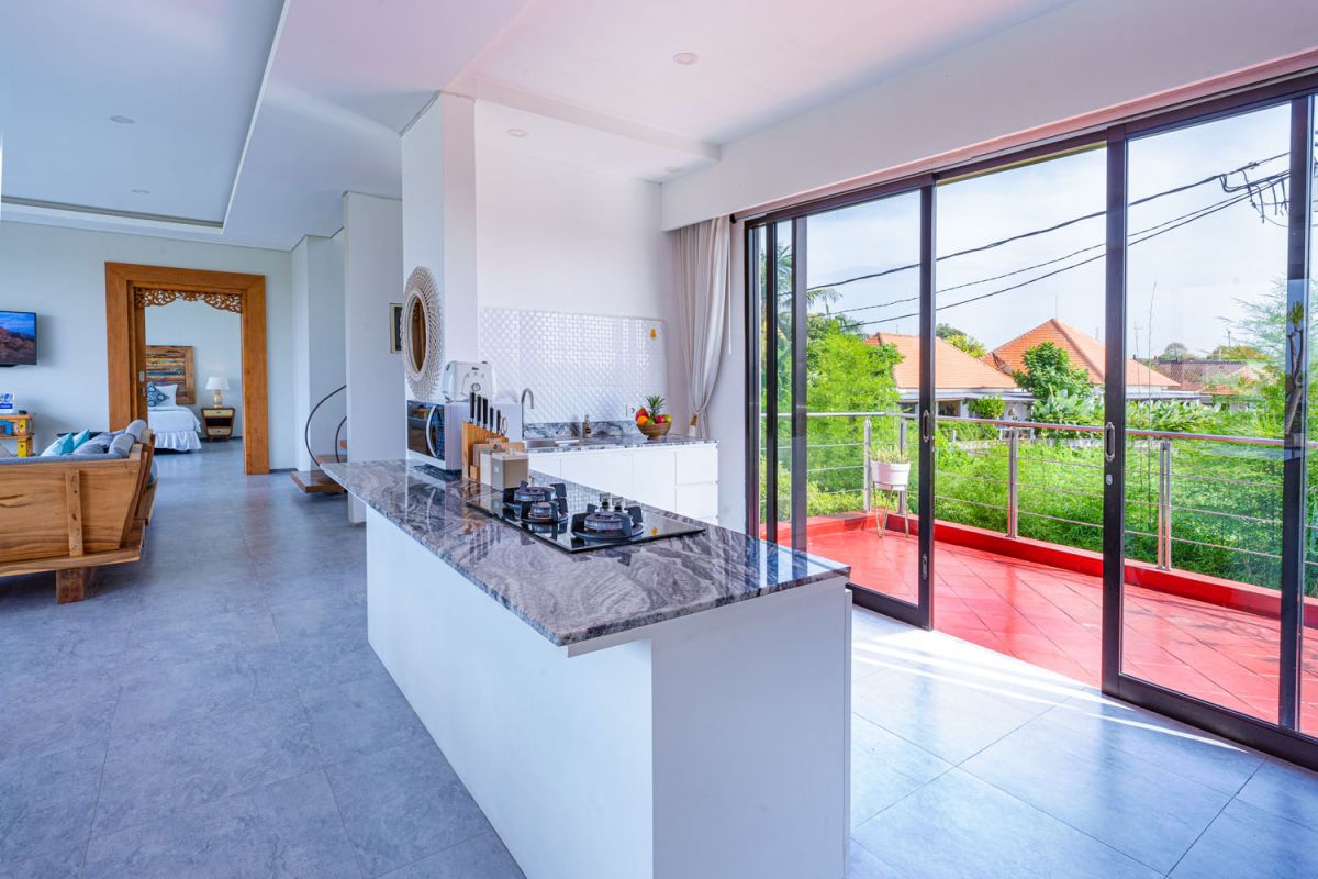 Spacious modern tropical dining room with wooden table, benches, and chairs, tropical flower centerpiece, open kitchen, and large windows overlooking lush greenery. - Bali Villas