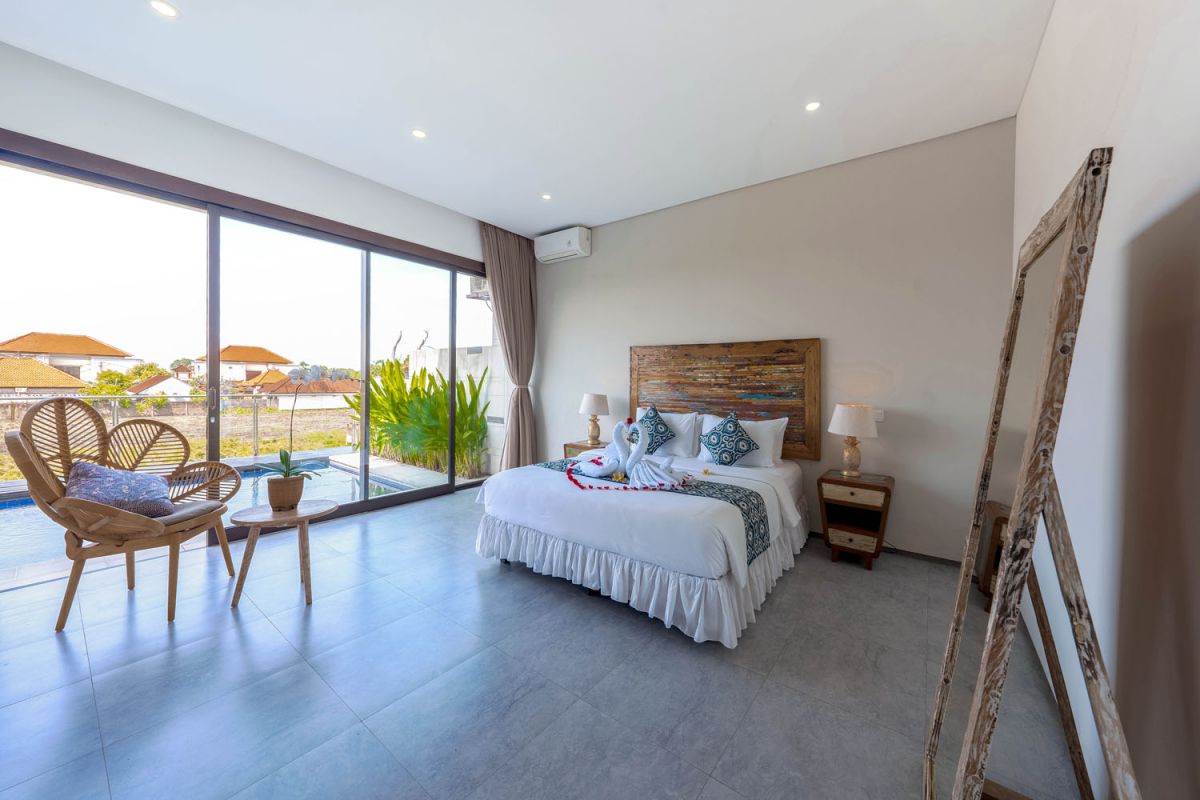 Spacious modern tropical dining room with wooden table, benches, and chairs, tropical flower centerpiece, open kitchen, and large windows overlooking lush greenery. - Bali Villas