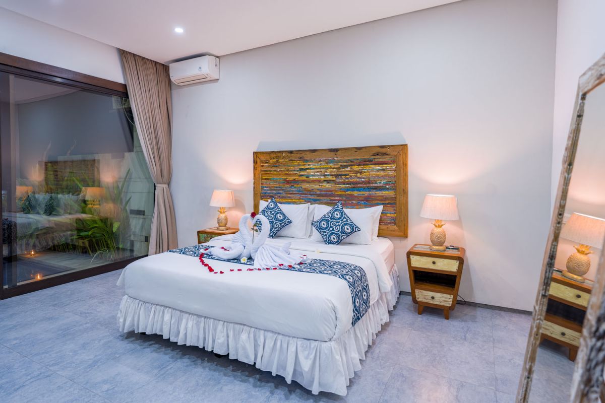 Spacious modern tropical dining room with wooden table, benches, and chairs, tropical flower centerpiece, open kitchen, and large windows overlooking lush greenery. - Bali Villas