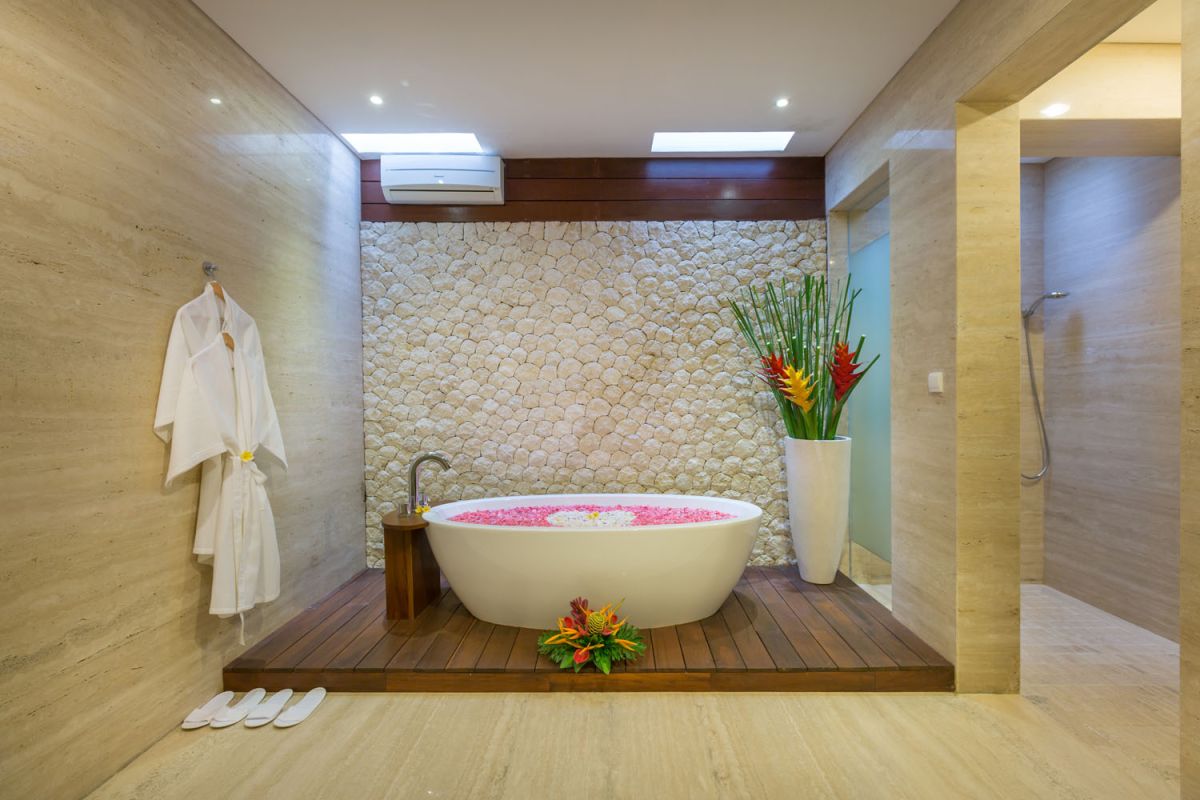 Luxurious spa bathroom with freestanding oval bathtub filled with pink water and rose petals, tropical bird-of-paradise flowers, white robe, and pebble wall accents. - Bali Villas