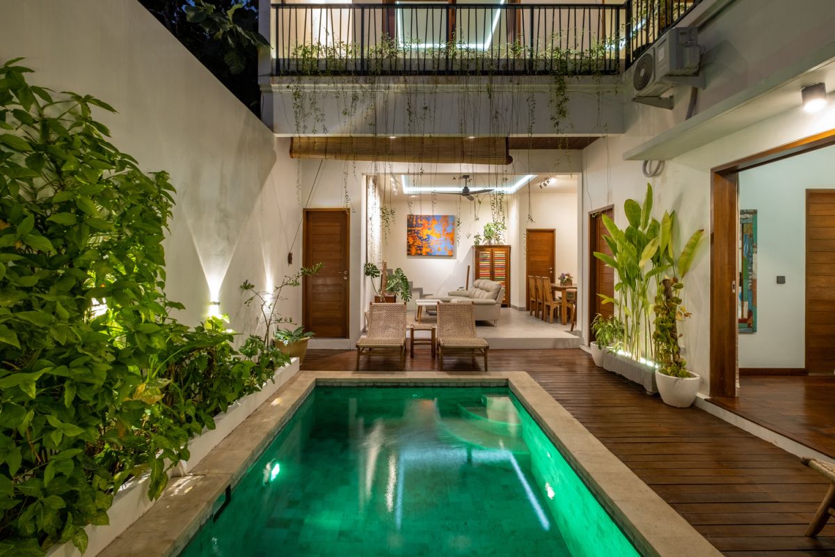 Overhead view of a private plunge pool in a tropical Bali villa, surrounded by wooden deck, lounge chairs, lush plants, and open bedroom doors. - Bali Villas