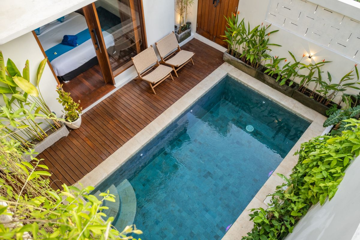 Overhead view of a private plunge pool in a tropical Bali villa, surrounded by wooden deck, lounge chairs, lush plants, and open bedroom doors. - Bali Villas