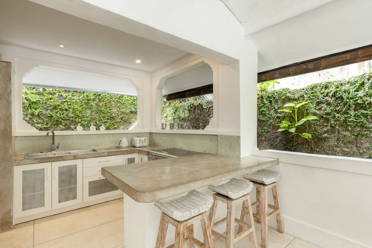 Modern white kitchen bar with concrete countertop, wooden stools, and lush tropical greenery outside arched windows. - Bali Villas