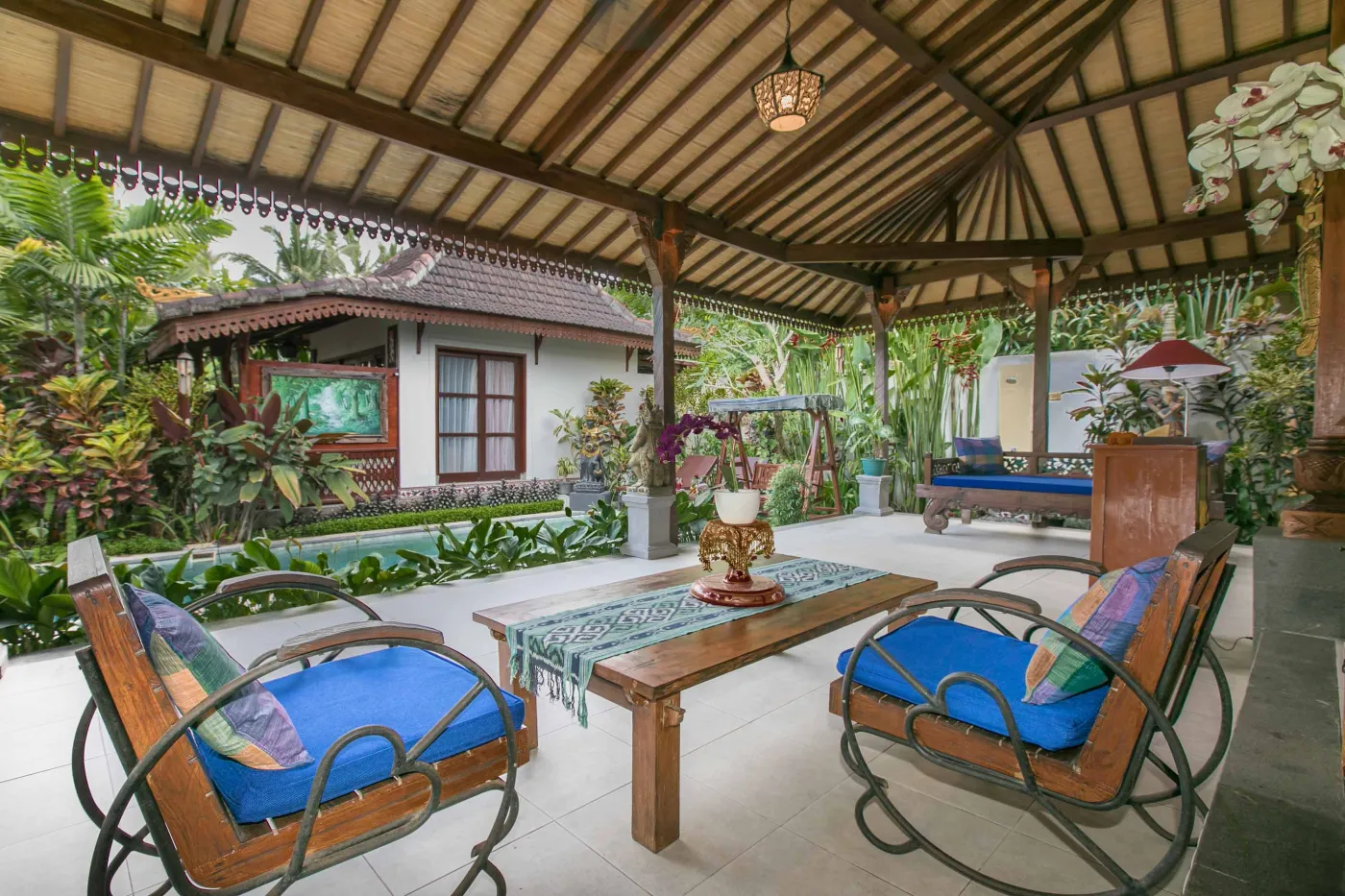 Spacious open-air Balinese pavilion with intricate carved wooden beams, hanging lanterns, rattan furniture, and tropical garden views. - Bali Villas