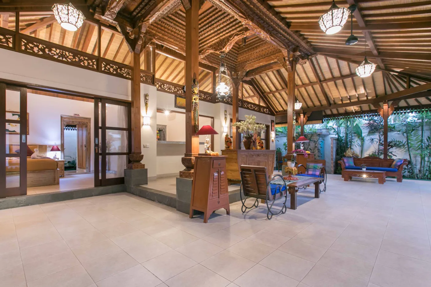 Spacious open-air Balinese pavilion with intricate carved wooden beams, hanging lanterns, rattan furniture, and tropical garden views. - Bali Villas