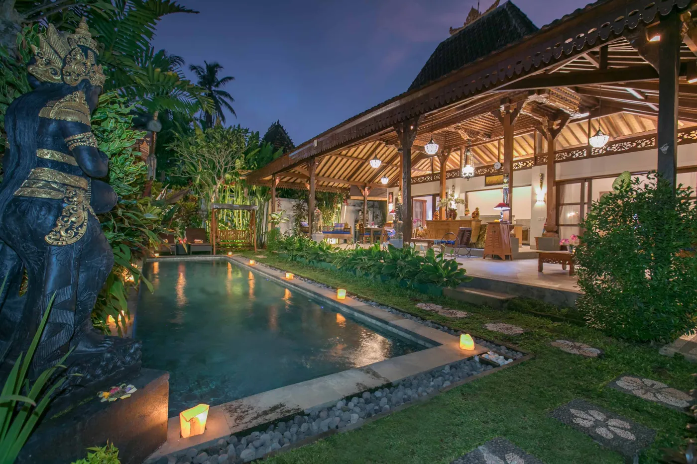 Spacious open-air Balinese pavilion with intricate carved wooden beams, hanging lanterns, rattan furniture, and tropical garden views. - Bali Villas