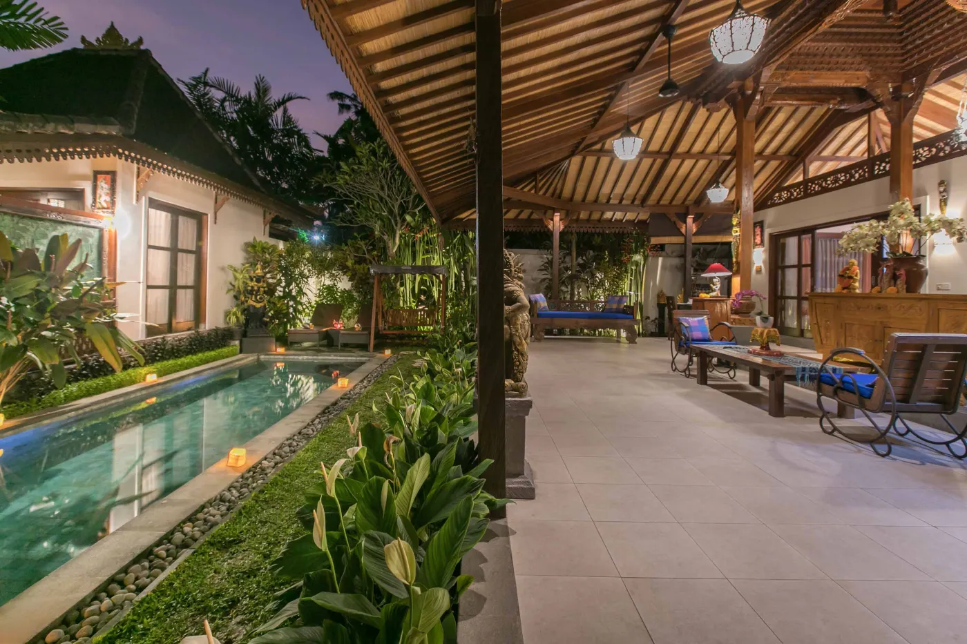 Spacious open-air Balinese pavilion with intricate carved wooden beams, hanging lanterns, rattan furniture, and tropical garden views. - Bali Villas