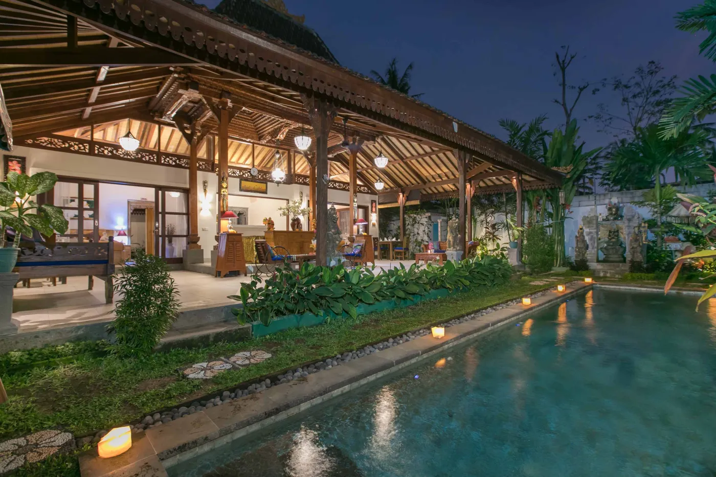 Spacious open-air Balinese pavilion with intricate carved wooden beams, hanging lanterns, rattan furniture, and tropical garden views. - Bali Villas