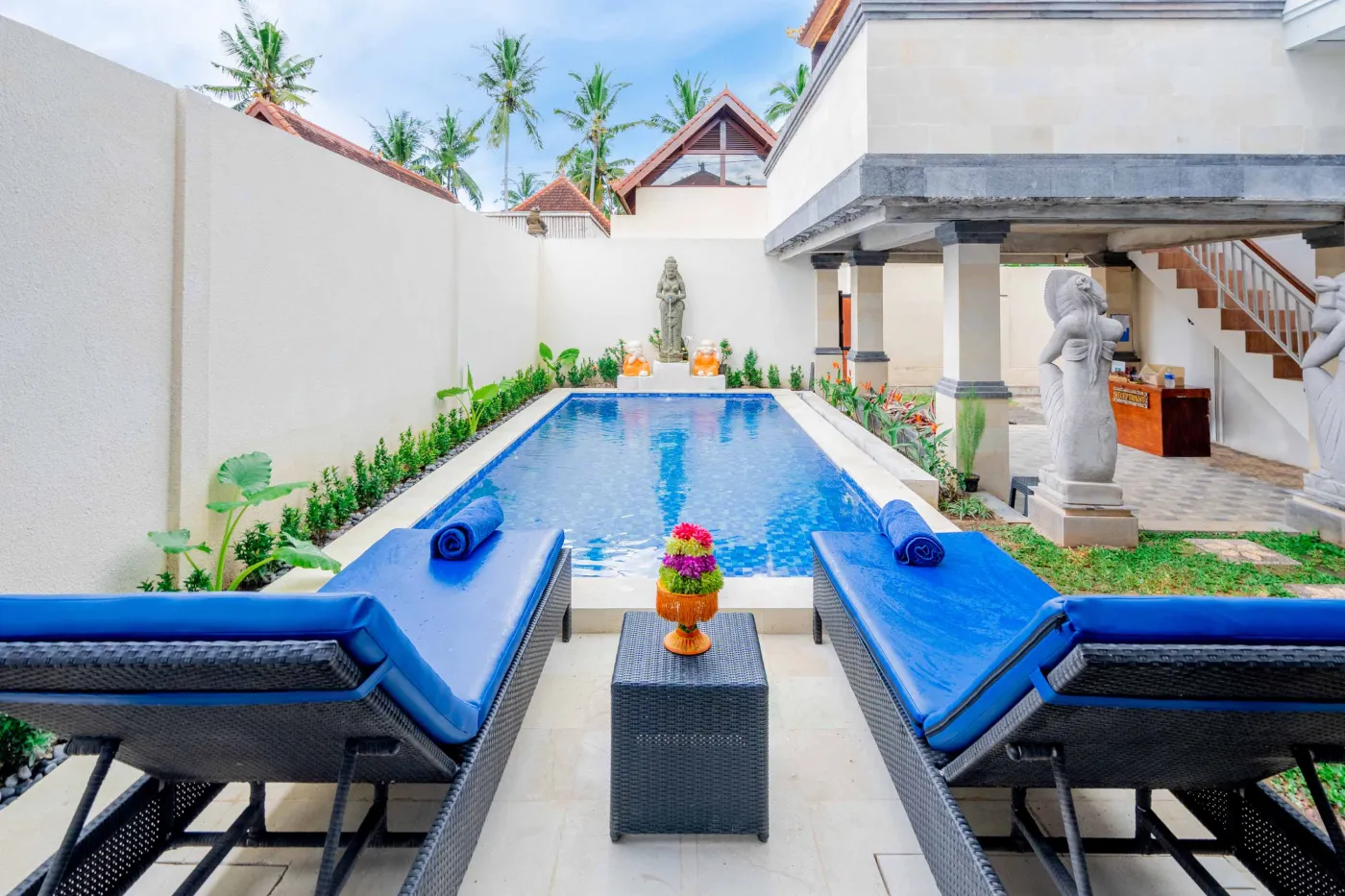 Serene blue infinity pool in a Balinese villa, surrounded by palm trees, stone guardian statues, lounge chairs, and tropical plants. - Bali Villas