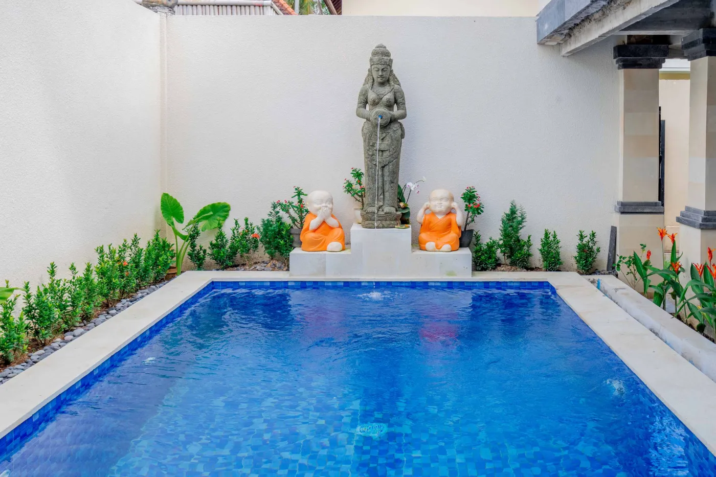 Serene Balinese villa courtyard with blue infinity pool, stone guardian statues, lounge chairs, tropical plants, and patterned tile patio. - Bali Villas