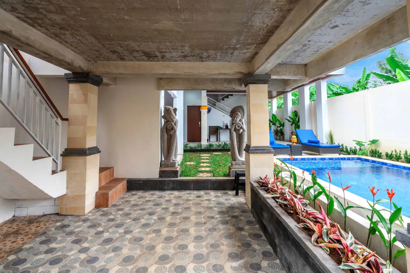 Serene Balinese villa courtyard with blue infinity pool, stone guardian statues, lounge chairs, tropical plants, and patterned tile patio. - Bali Villas