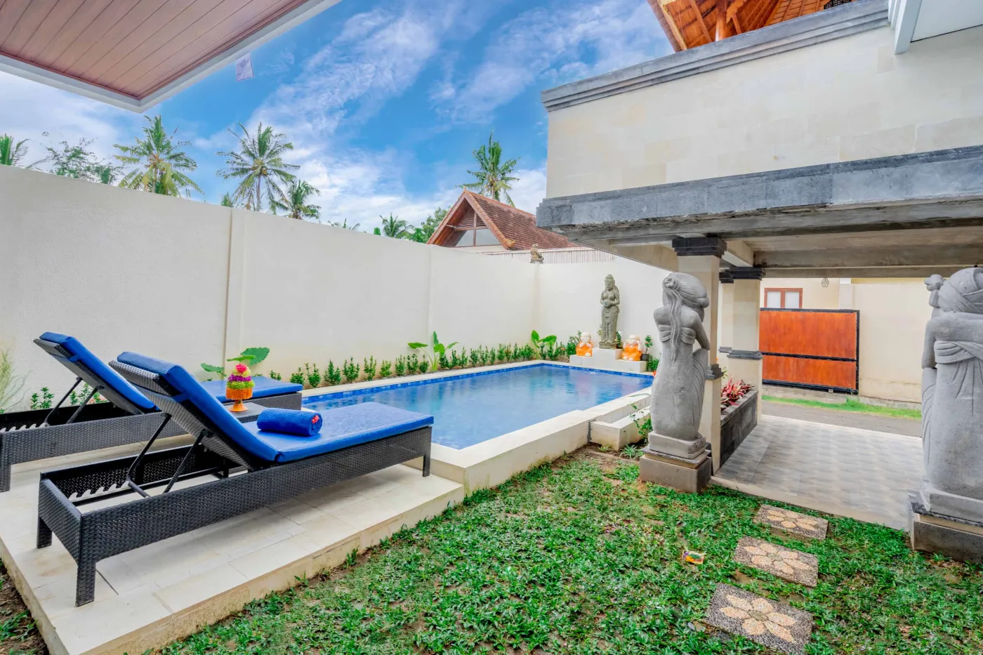 Serene Balinese villa courtyard with blue infinity pool, stone guardian statues, lounge chairs, tropical plants, and patterned tile patio. - Bali Villas