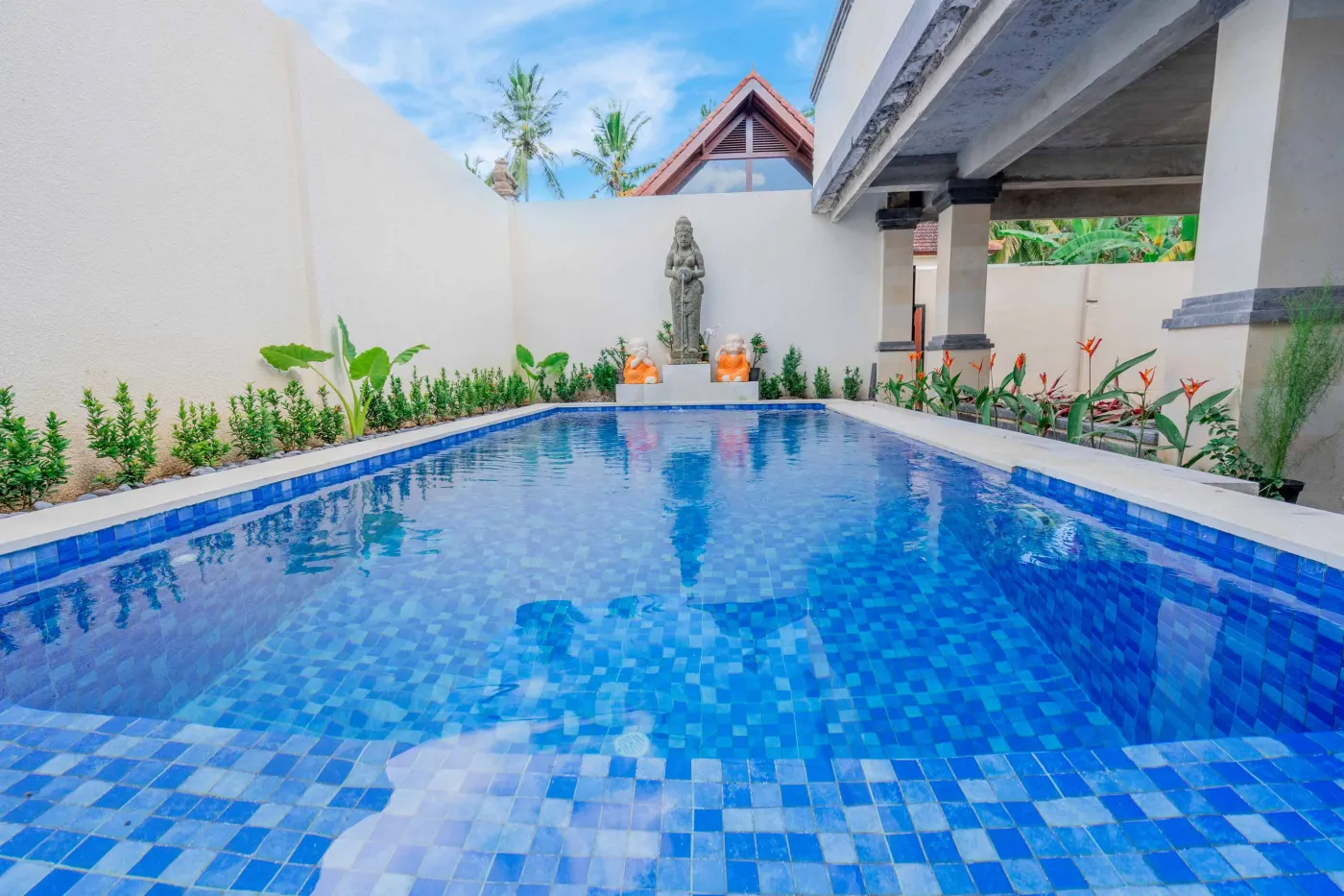 Serene Balinese villa courtyard with blue infinity pool, stone guardian statues, lounge chairs, tropical plants, and patterned tile patio. - Bali Villas