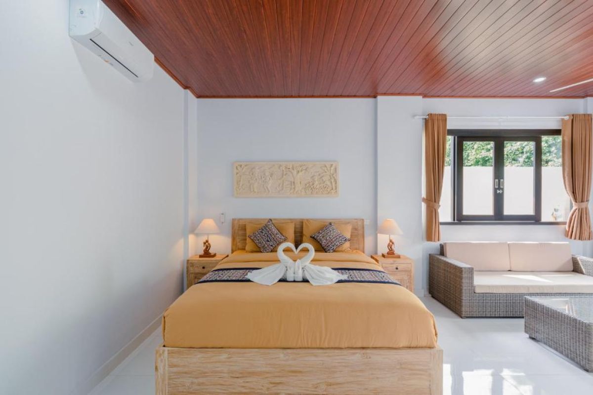 Spacious Balinese-style hotel bedroom with wooden ceiling fan, king bed, wall-mounted TV, and orange curtains. - Bali Villas