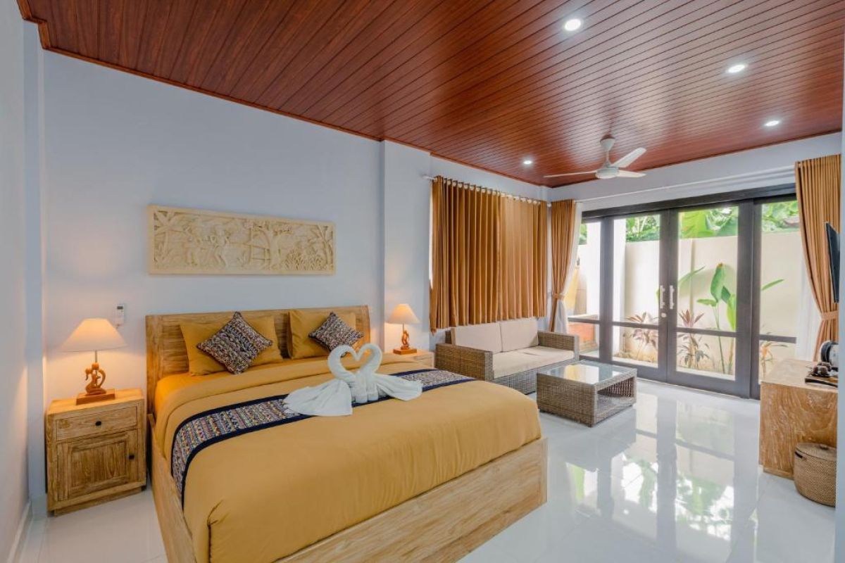 Spacious Balinese-style hotel bedroom with wooden ceiling fan, king bed, wall-mounted TV, and orange curtains. - Bali Villas