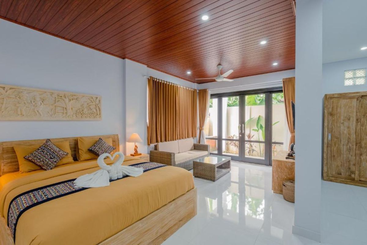 Spacious Balinese-style hotel bedroom with wooden ceiling fan, king bed, wall-mounted TV, and orange curtains. - Bali Villas
