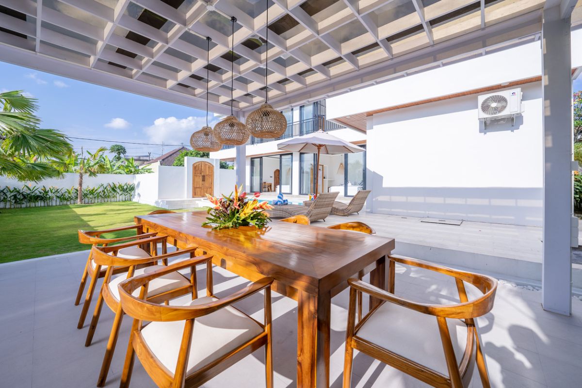 Modern tropical kitchen with curved white counter, rattan bar stools, hanging woven lamps, abstract wall art, and lush green views through large windows. - Bali Villas