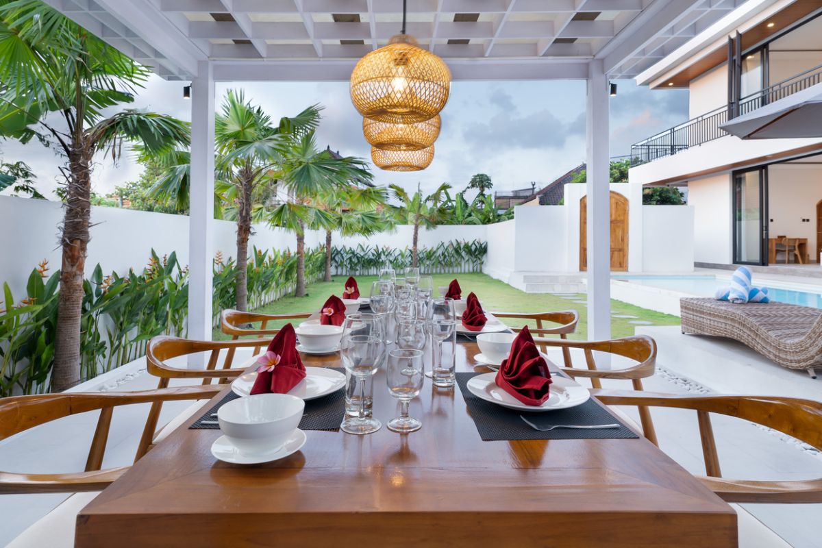 Modern tropical kitchen with curved white counter, rattan bar stools, hanging woven lamps, abstract wall art, and lush green views through large windows. - Bali Villas