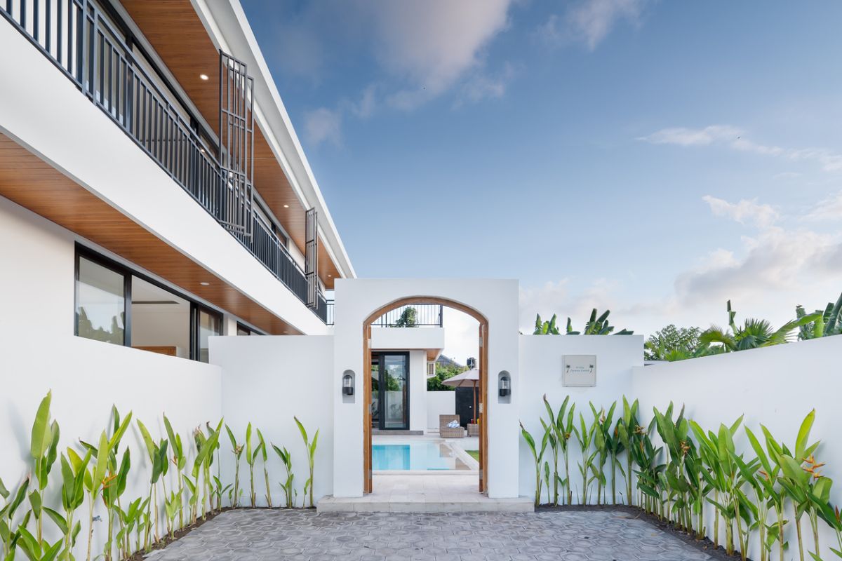 Modern tropical kitchen with curved white counter, rattan bar stools, hanging woven lamps, abstract wall art, and lush green views through large windows. - Bali Villas
