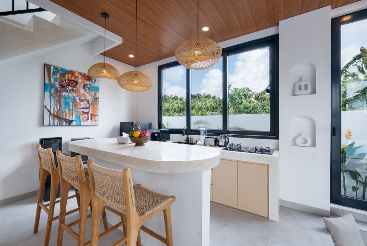 Modern tropical kitchen with curved white counter, rattan bar stools, hanging woven lamps, abstract wall art, and lush green views through large windows. - Bali Villas