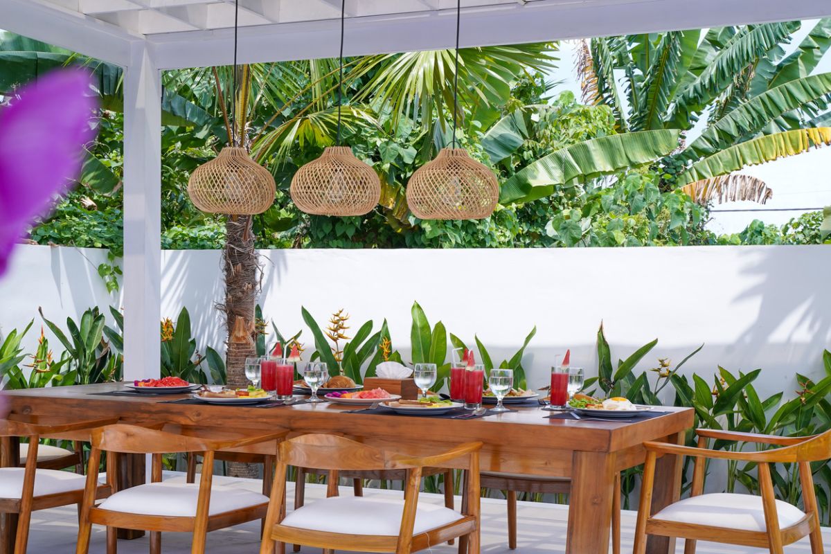 Modern tropical kitchen with curved white counter, rattan bar stools, hanging woven lamps, abstract wall art, and lush green views through large windows. - Bali Villas