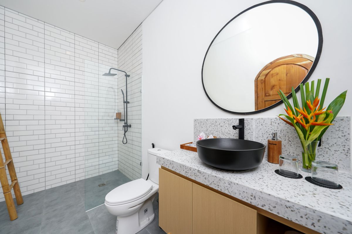 Modern tropical kitchen with curved white counter, rattan bar stools, hanging woven lamps, abstract wall art, and lush green views through large windows. - Bali Villas